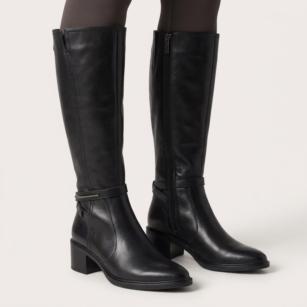 Pormenor de um par de botas Carmela pretas de cano alto em pele, com collants escuros sobre fundo claro de estúdio