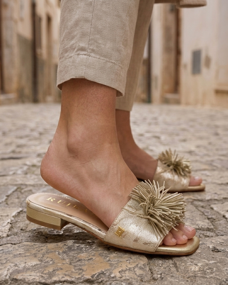 Vista lateral aproximada de chinelo RUIKA metalizado champanhe/platino com pompom de franjas e monograma dourado "R" cravado na gáspea, calçado sobre calçada de pedra irregular com calças de linho bege