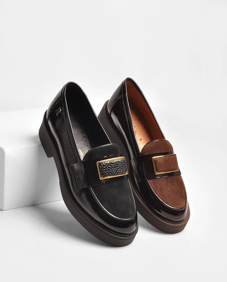 Par de mocassins Ruika em pele envernizada, um preto e outro castanho, com espelho em camurça e fivela dourada, apoiados num bloco branco