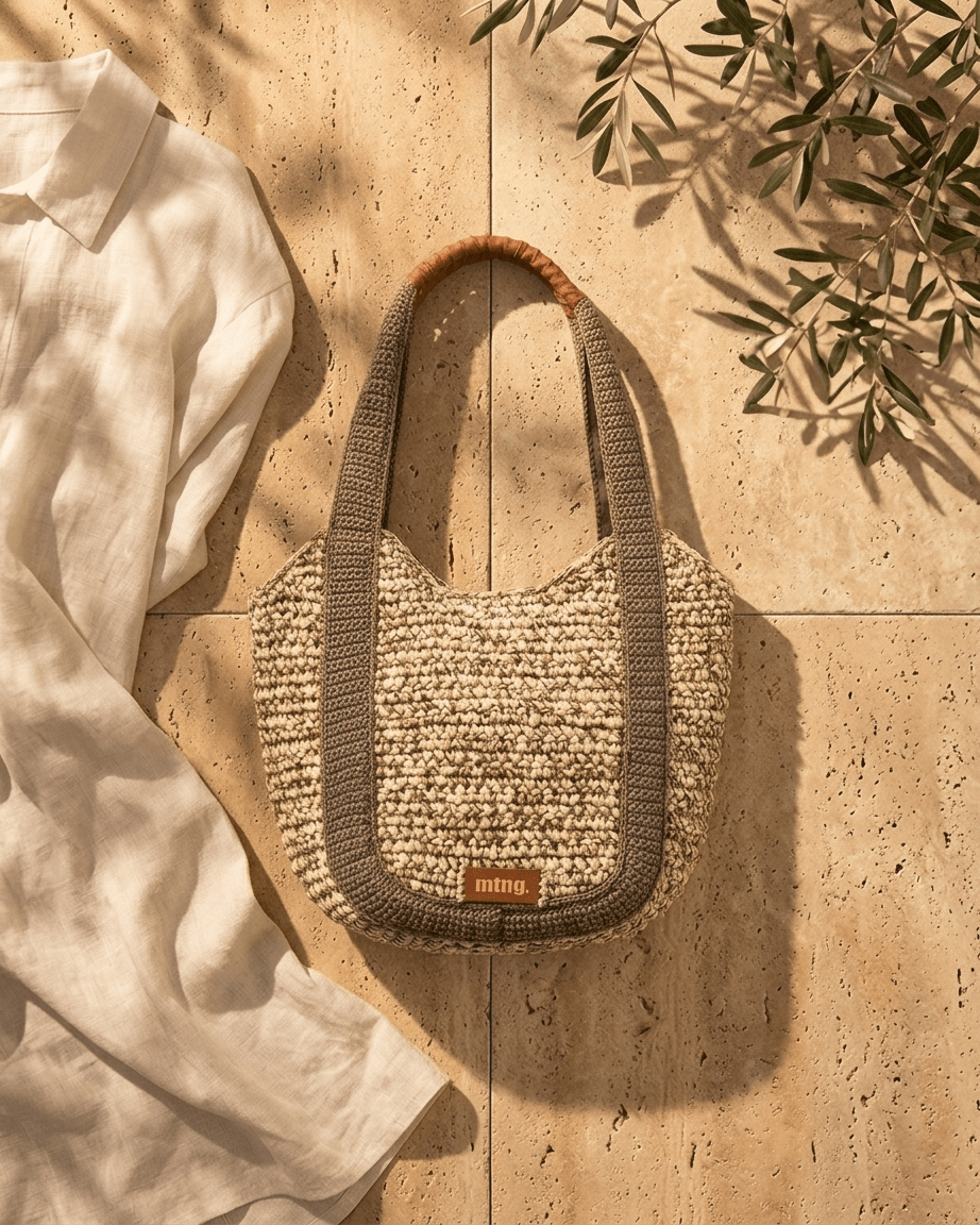 Vista superior da bolsa shopper Mustang Mar em ráfia de papel creme com bordas em macramê cáqui e alças com acabamento em couro castanho, sobre pavimento de travertino com ramo de oliveira