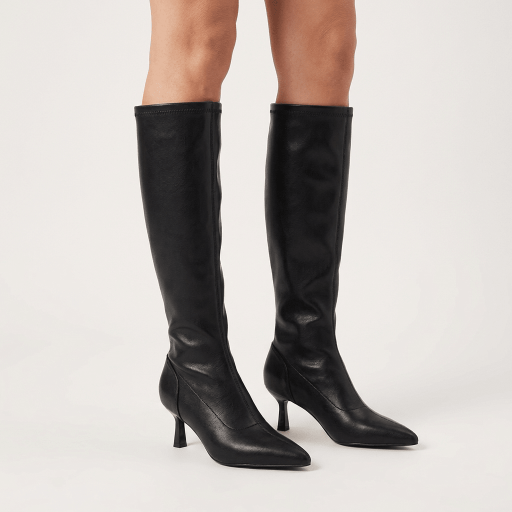 Pernas femininas com par de botas de cano alto Mariamare pretas até ao joelho, bico fino e salto fino, em fundo neutro claro