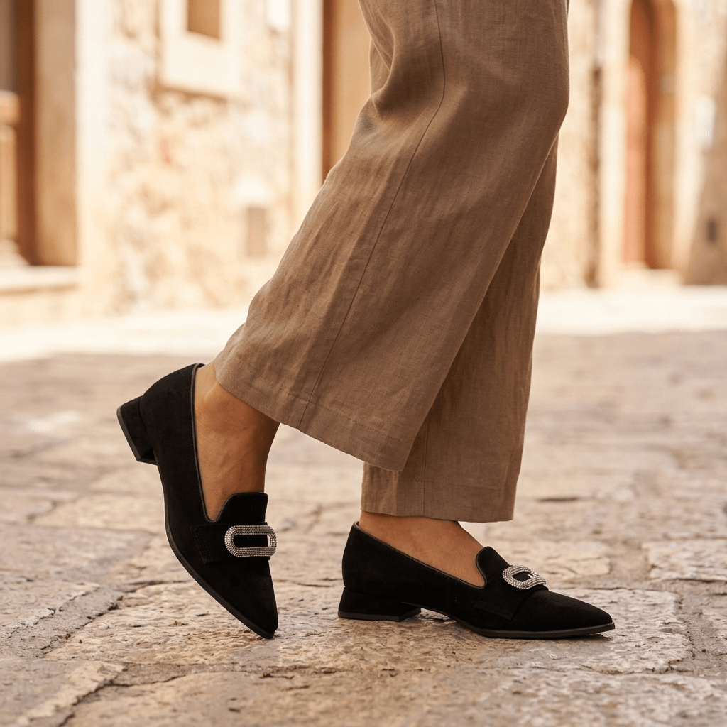Pés femininos com sapatos Turtle de camurça preta e aplicação decorativa prateada, a caminhar em calçada de pedra com calças de linho