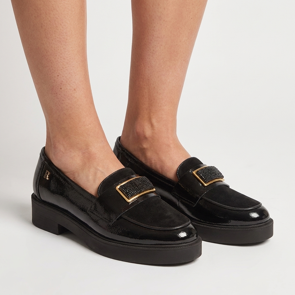 Pés de mulher a calçar mocassins Ruika pretos em verniz com fivela dourada e sola robusta, sobre fundo claro