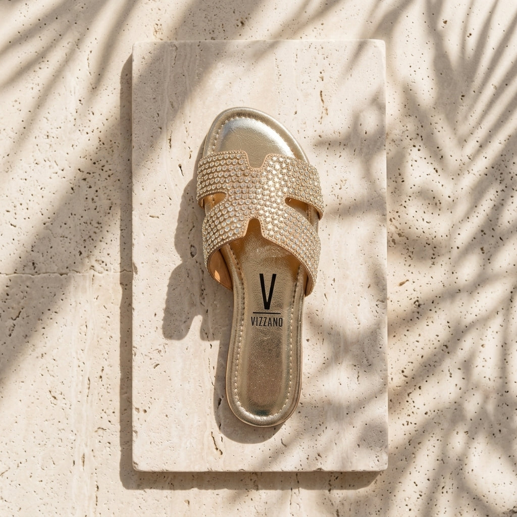 Detalhe do chinelo raso Vizzano dourado com pedrarias e logótipo na palmilha, fotografado contra parede com sombra de folhagem