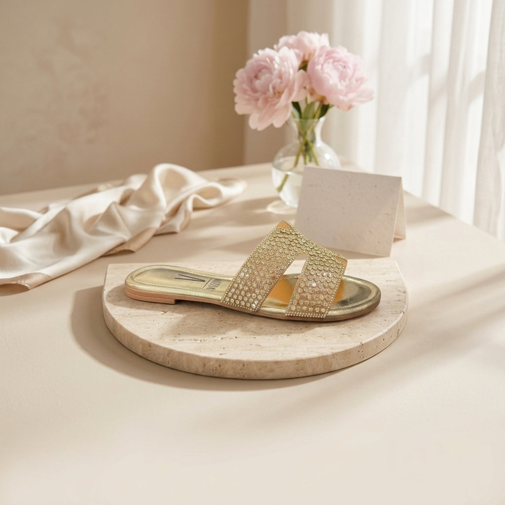Chinelo raso Vizzano dourado com pedrarias em composição de still life, junto a flores cor-de-rosa e tecido claro sobre superfície de pedra