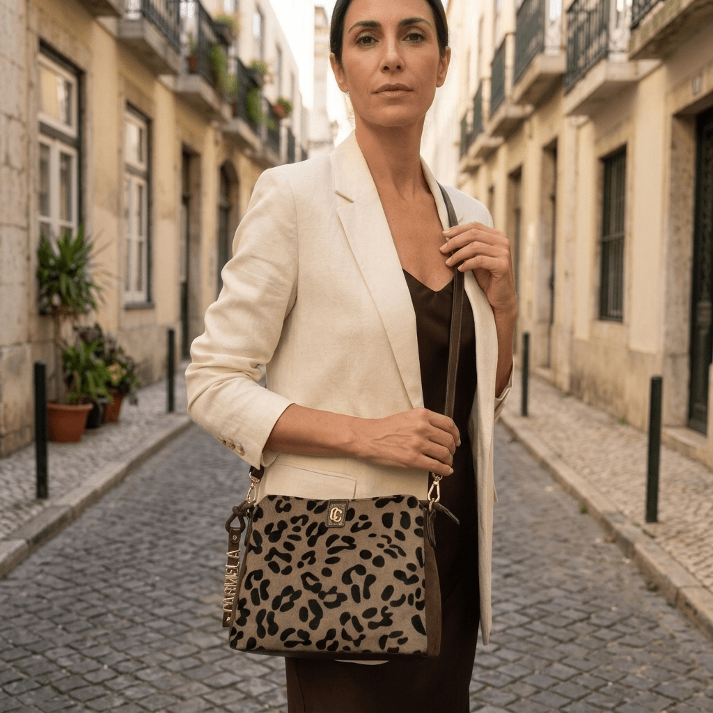 Bolsa Carmela camurça taupe usada a tiracolo por uma mulher de blazer creme, numa rua estreita de Lisboa.