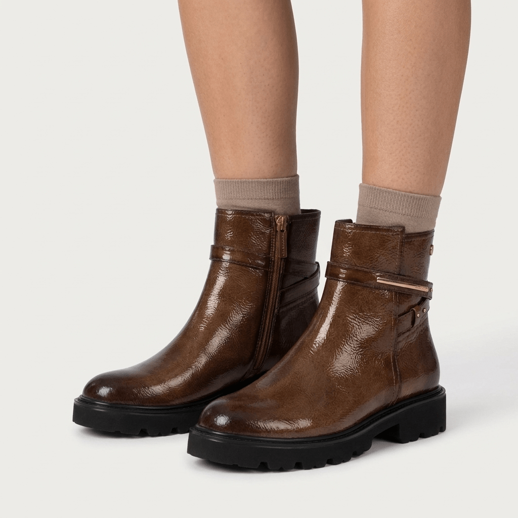 Par de botins Carmela castanho bronzeado em verniz, calçados com meias beges sobre fundo cinzento claro