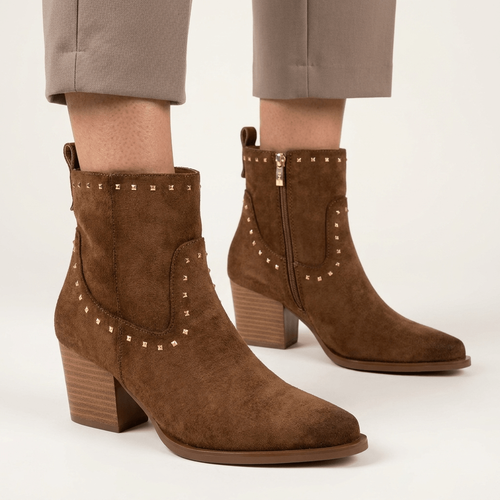 Pernas de mulher com calças cinzentas a calçar botins XTi camel em antelina com apliques metálicos e fecho lateral, em estúdio