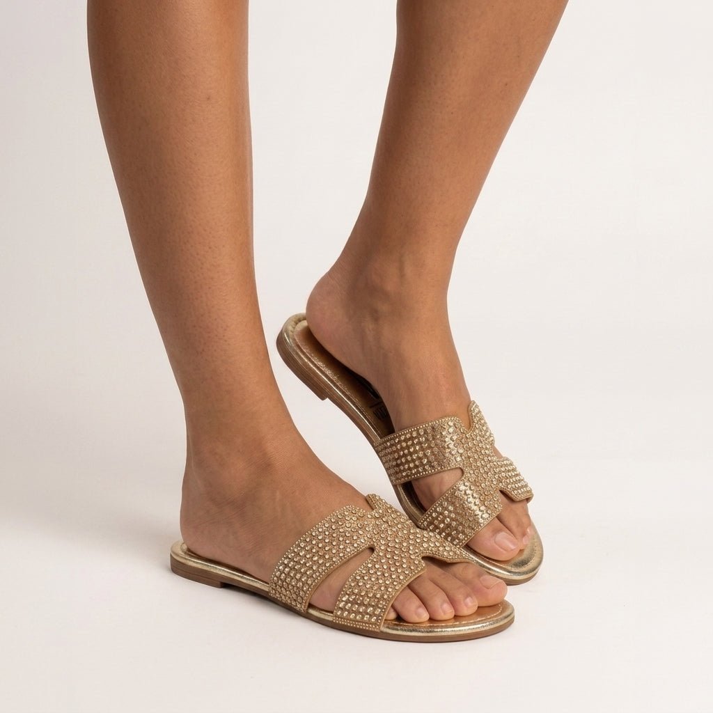 Detalhe dos pés calçados com o chinelo raso Vizzano dourado com pedrarias, em pé sobre piso claro em tom natural