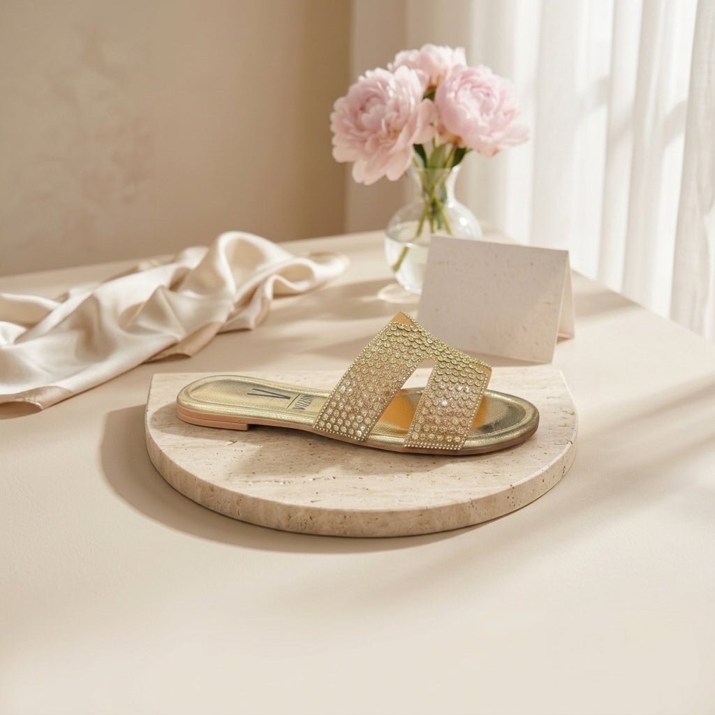 Chinelo raso Vizzano dourado com pedrarias em composição de still life, junto a flores cor-de-rosa e tecido claro sobre superfície de pedra