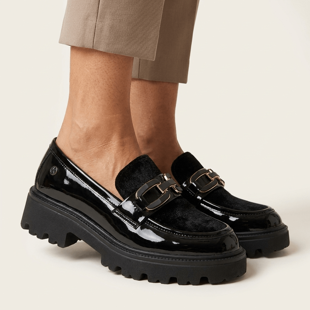 Mocassim Keys preto em verniz, visto de três quartos, com tira aveludada, aplique metálico e sola tratorada, calçado com calças bege em fundo claro