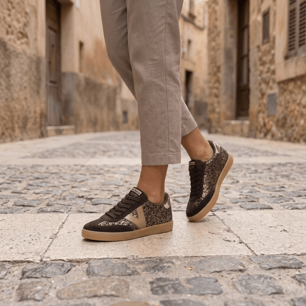 Pernas com calças taupe a caminhar com sapatilhas Victória Berlín Glitter castanho numa rua de pedra