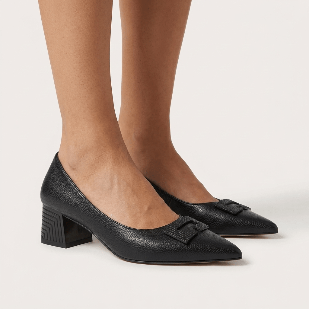 Pés femininos com sapatos clássicos Azarey pretos, bico fino, tacão geométrico ranhurado e adorno no peito do pé, fundo claro