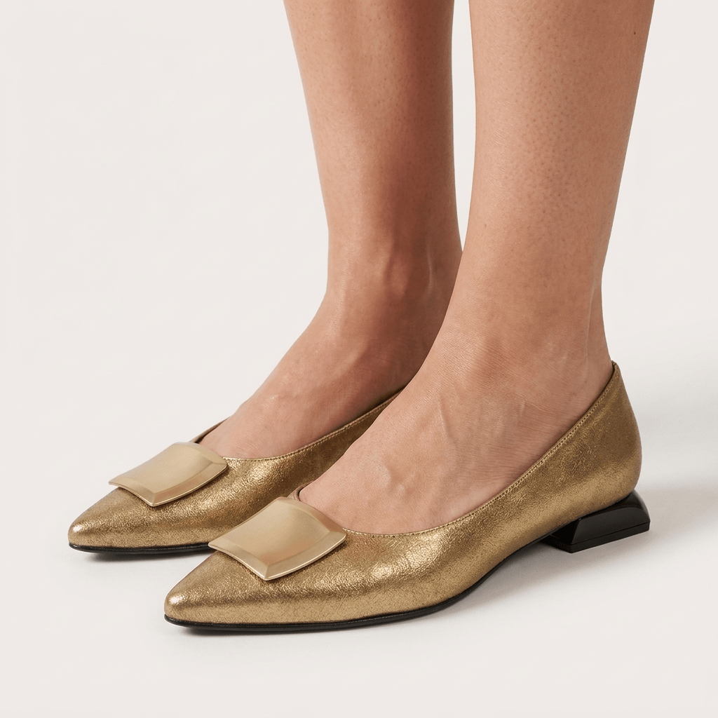 Pés de mulher com sapatos Daniella Vega dourados de bico fino e aplique metálico, em vista frontal sobre fundo claro
